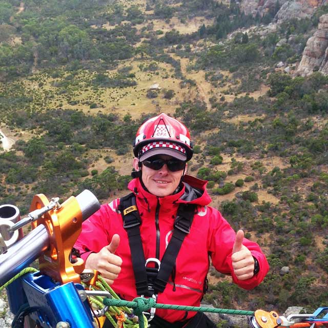 Man wearing red clothing and safety gear against a distant background, suspended from a high place by abseiling equipment