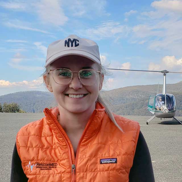 Woman wearing white cap and orange medical vest stands in front of a rescue helicopter
