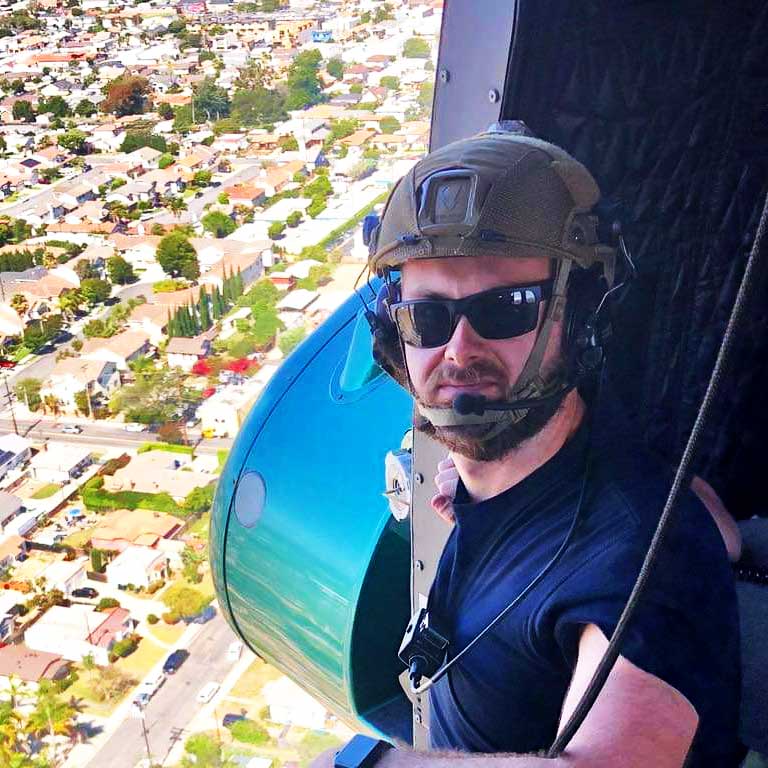 Man wearing aviators helmet and sunglasses next to window of helicopter flying over an urban area
